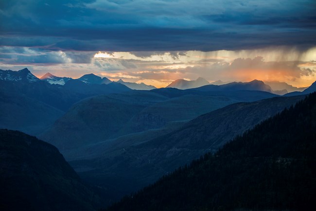 A range of mountains which look blue beneath an orange sunset and stormy sky.