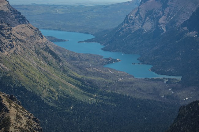 A blue lake trails behind the curve of a mountain in the foreground. Mountains on the other side similarly slope up forming a "u" shape with the lake in the middle of both ranges.