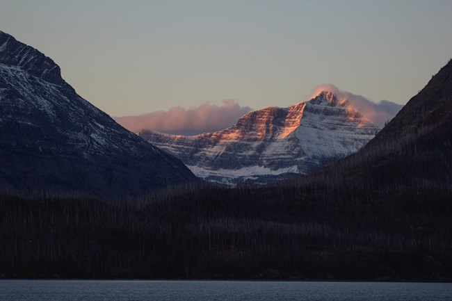 Two mountain peaks partially covered in clouds and coated in snow. They are tinged pink from the sunset under a blue and yellow sky.