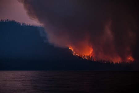 A photo taken as night is falling of a wildfire burning a steep slope. The fire glows orange and dark smoke billows into the sky.