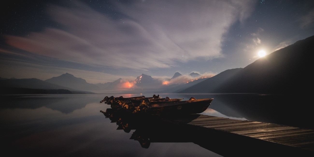 A glassy lake on a moonlit night, with a boat dock and boats in the foreground, and a wildfire glowing in mountains in the background.