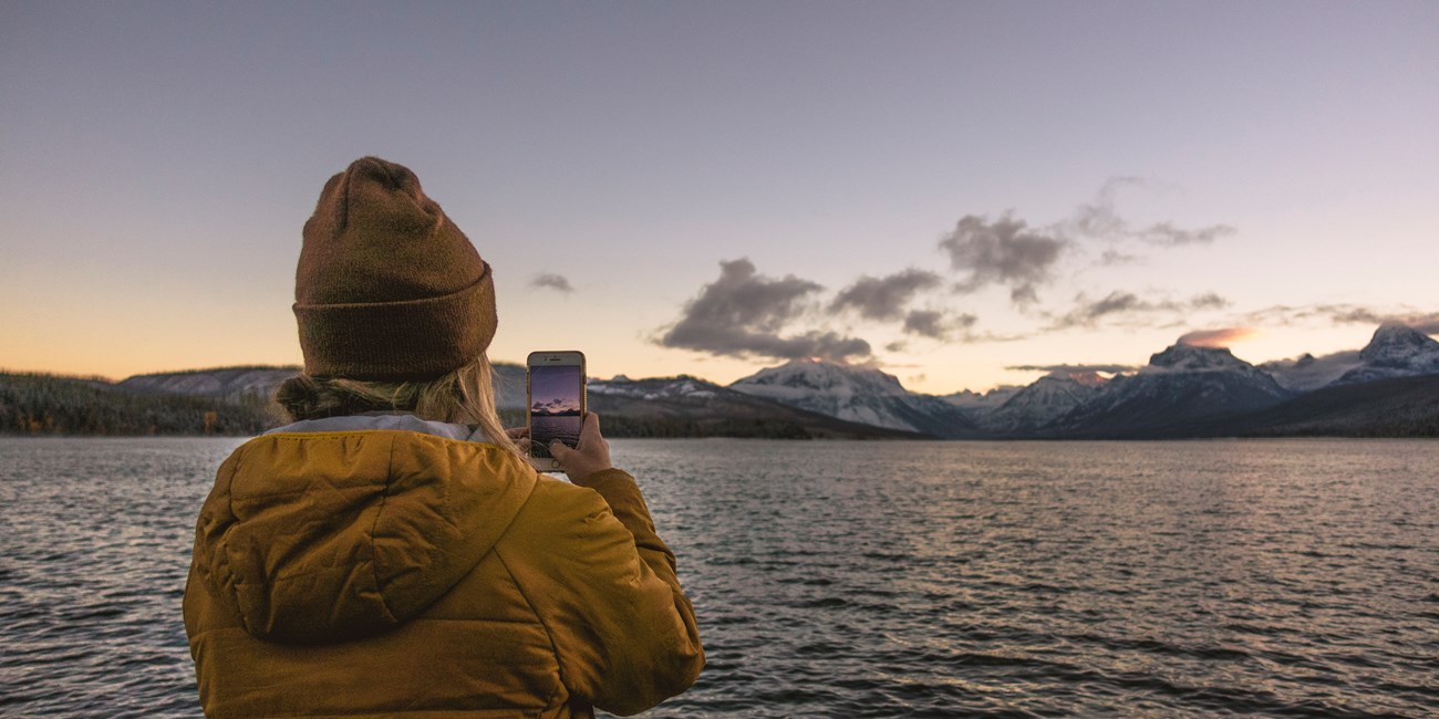 A person in a coat and hat photographs sunrise over mountains with a phone with a lake in the foreground.