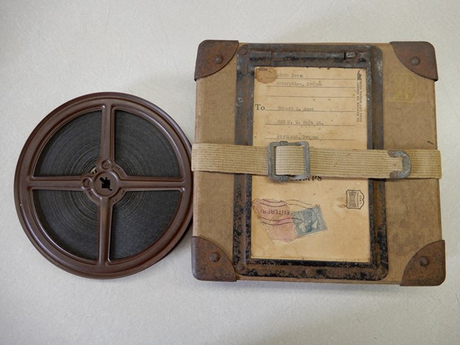 A reel of physical film in a metal container sits next to a canvas and metal box used to mail it in.