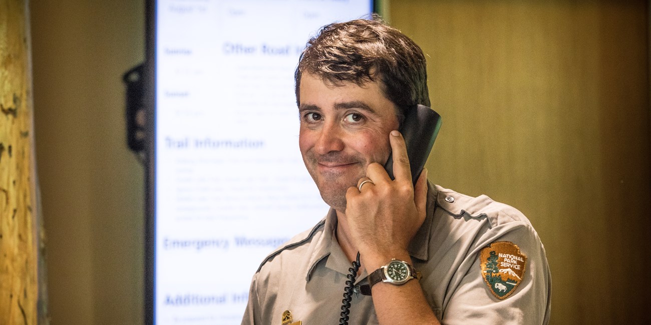 A park ranger smiles at the camera while holding a phone up to their ear.