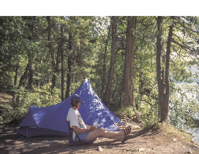 A camper sits in front of a blue tent facing a lake. Behind the tent is a very green forest.