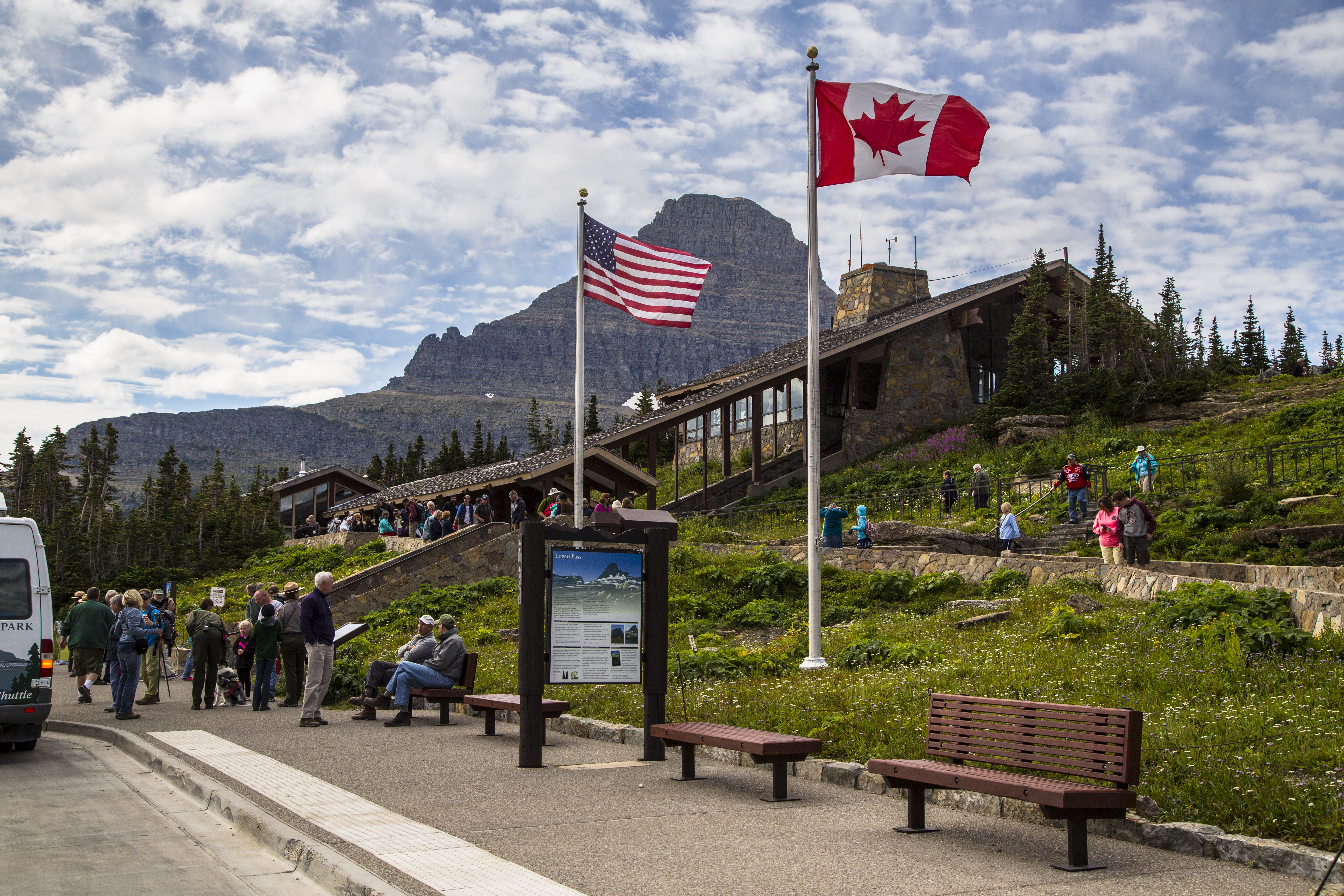 Visitors mill around the Logan Pass shuttle stop and Logan Pass Visitor Center. A tall peak looms in the background.