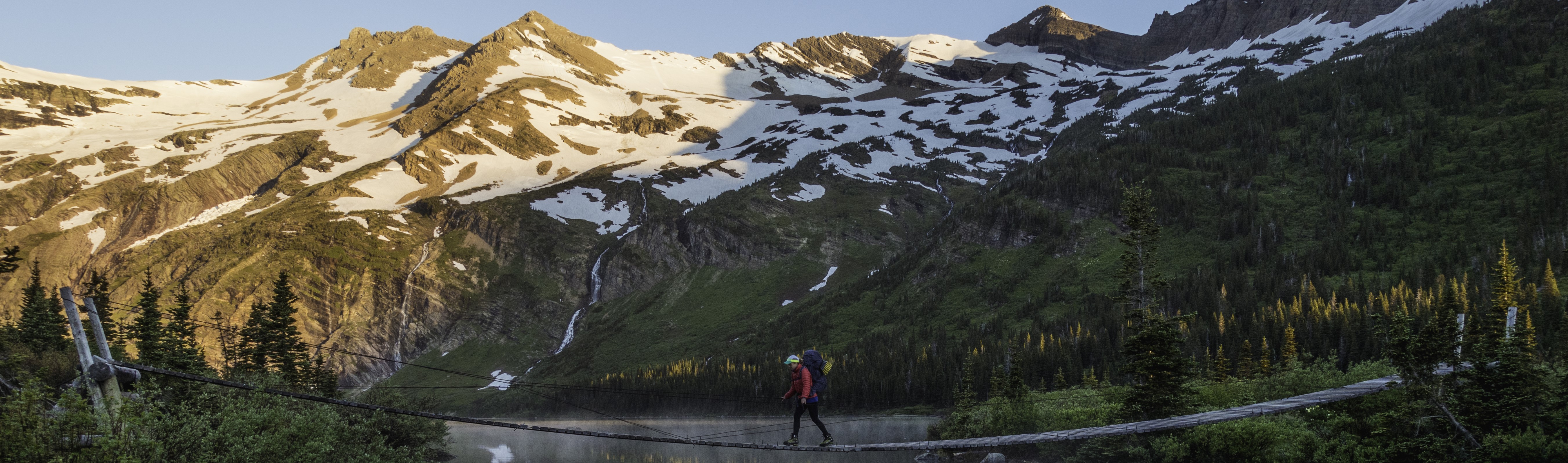 A backpacker crosses a suspension bridge in front of snow covered mountains.