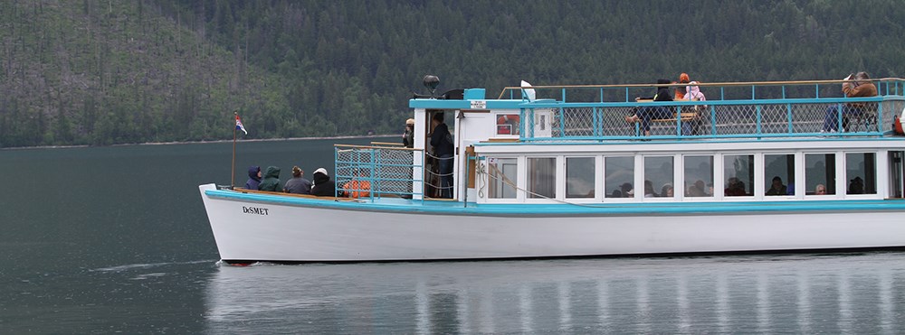 large wooden boat cruises lake with visitors on top deck, bow, and in cabin