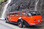 Red Bus with black top drives past waterfall