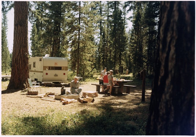 A family of four is in a campground. The father is building a fire and the two young children are at the picnic table with their mother.