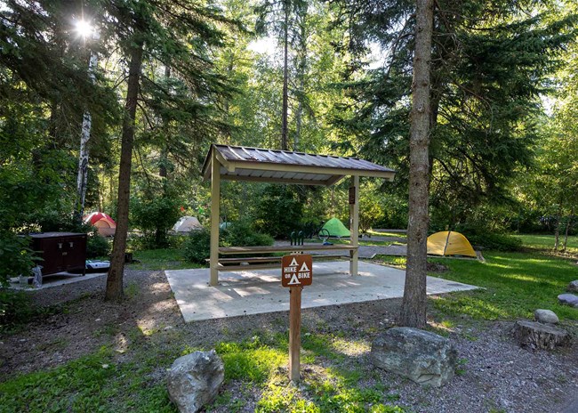 A wooden sign indicates the campsite is for hiker/bikers. Behind it are colorful tents seen throughout the woods.