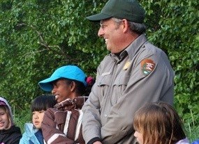 a park ranger smiles and holds the hands of several children during an outdoor activity