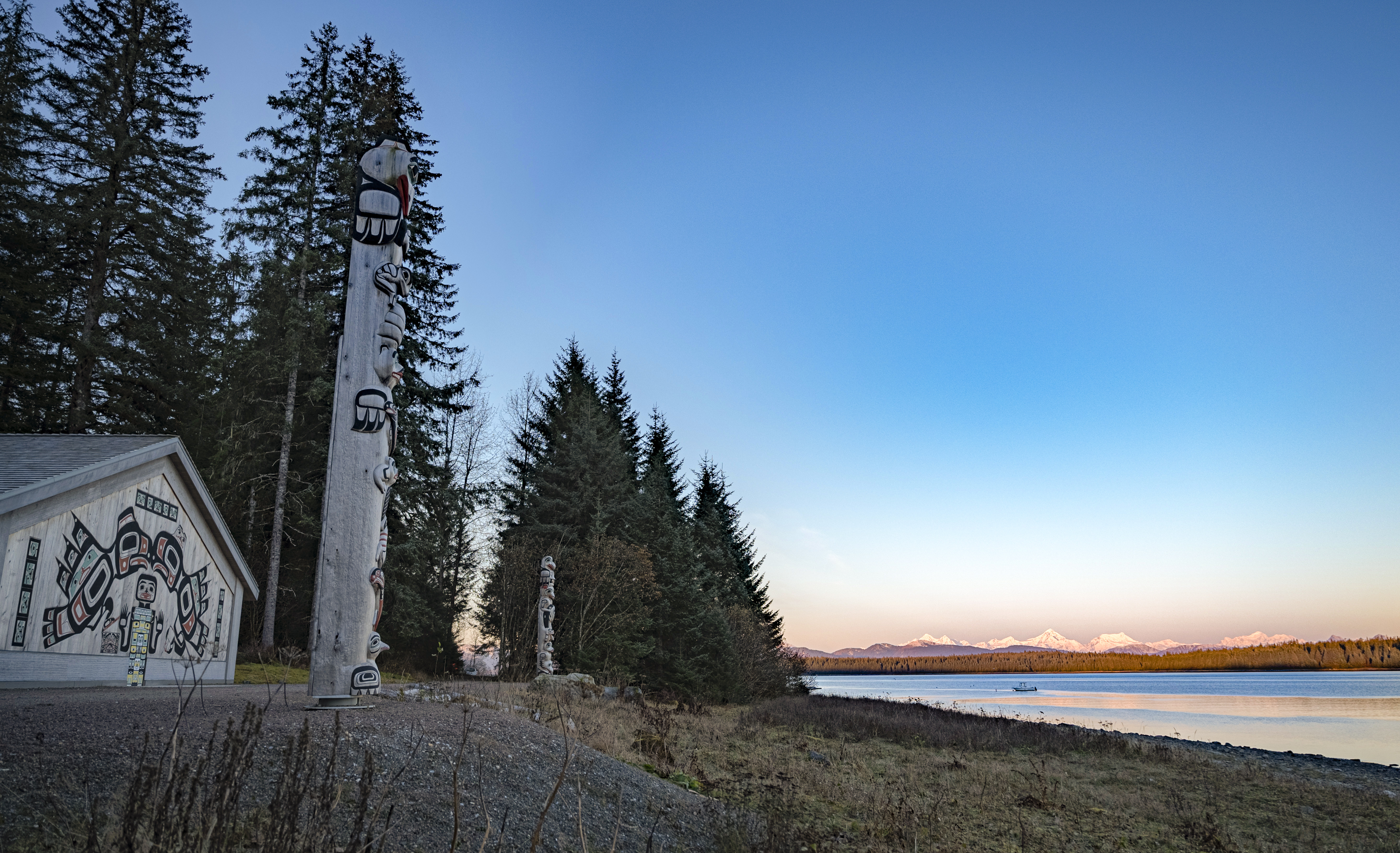 Two totem poles stand in front of the Huna Tribal house, during sunrise on a clear day. Distant mountains glow pink/orange.