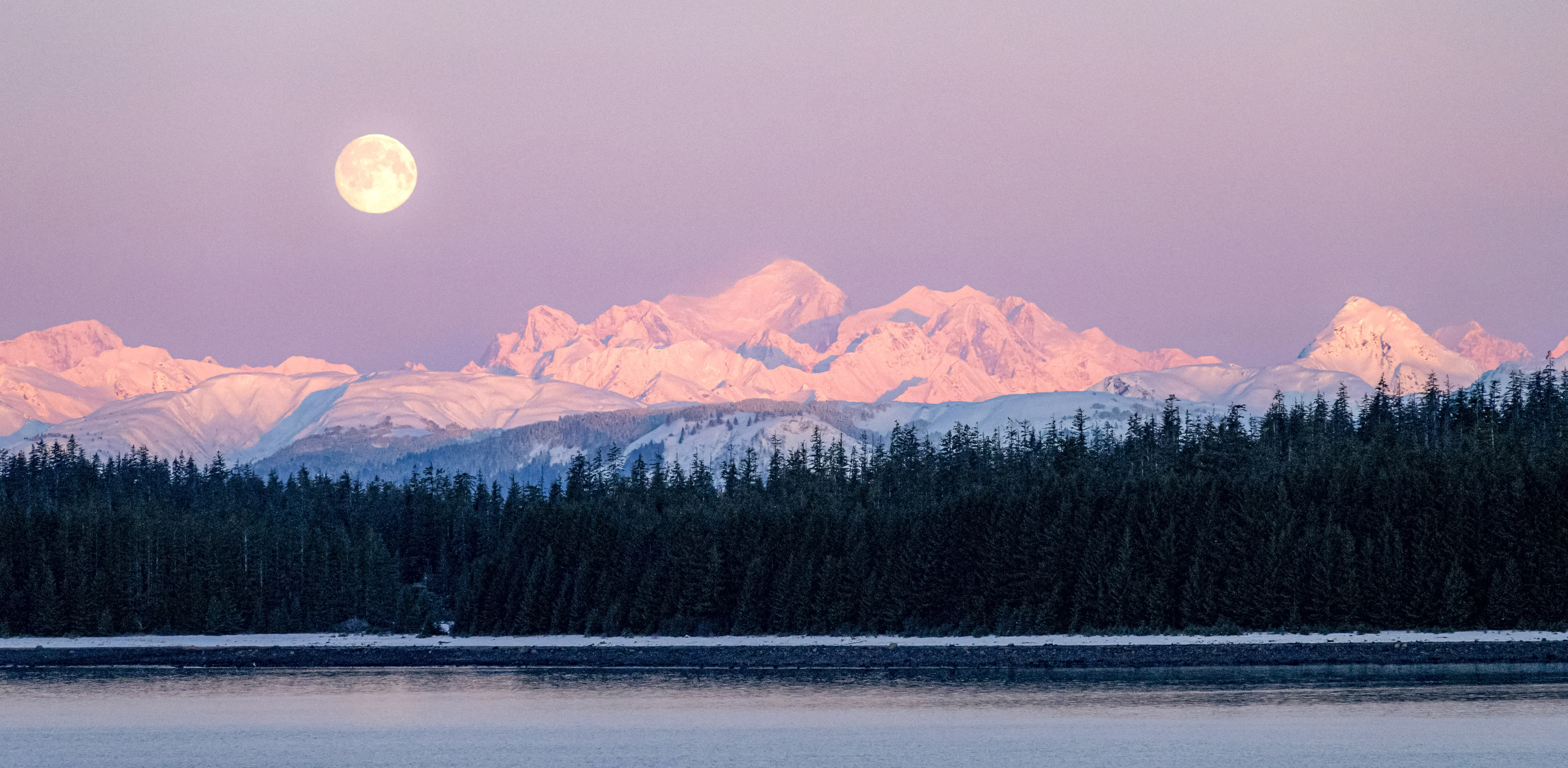 A bright full moon sits to the left above Mt Fairweather, snow colored pink by alpenglow. Bartlett Cove in foreground.