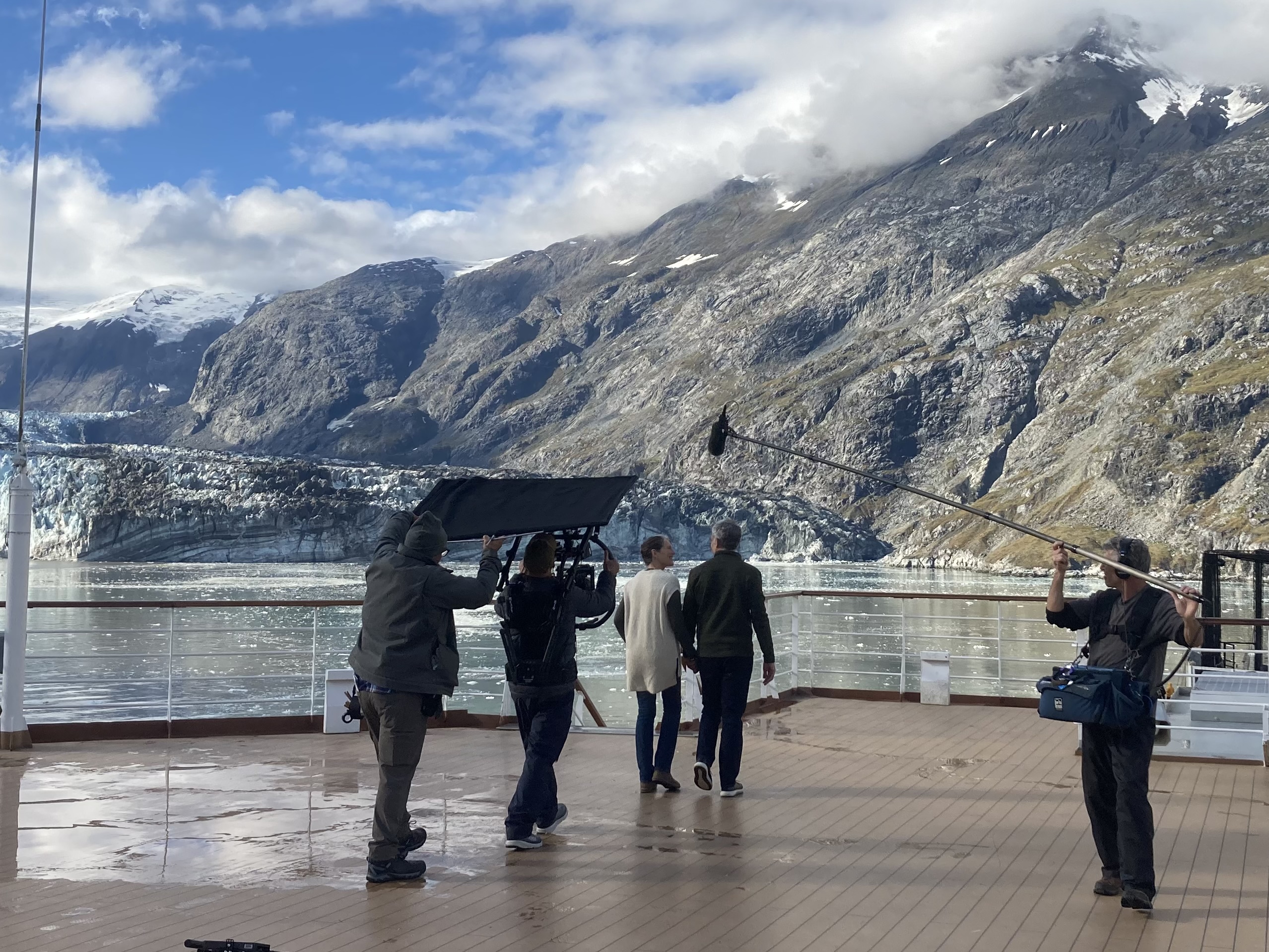 A male actor and afemale actor walk hand in hand, while being filmed by a crew of three people. THey are on the deck of a cruise ship in front of a glacier with grey, snow peaked mountains in the background..