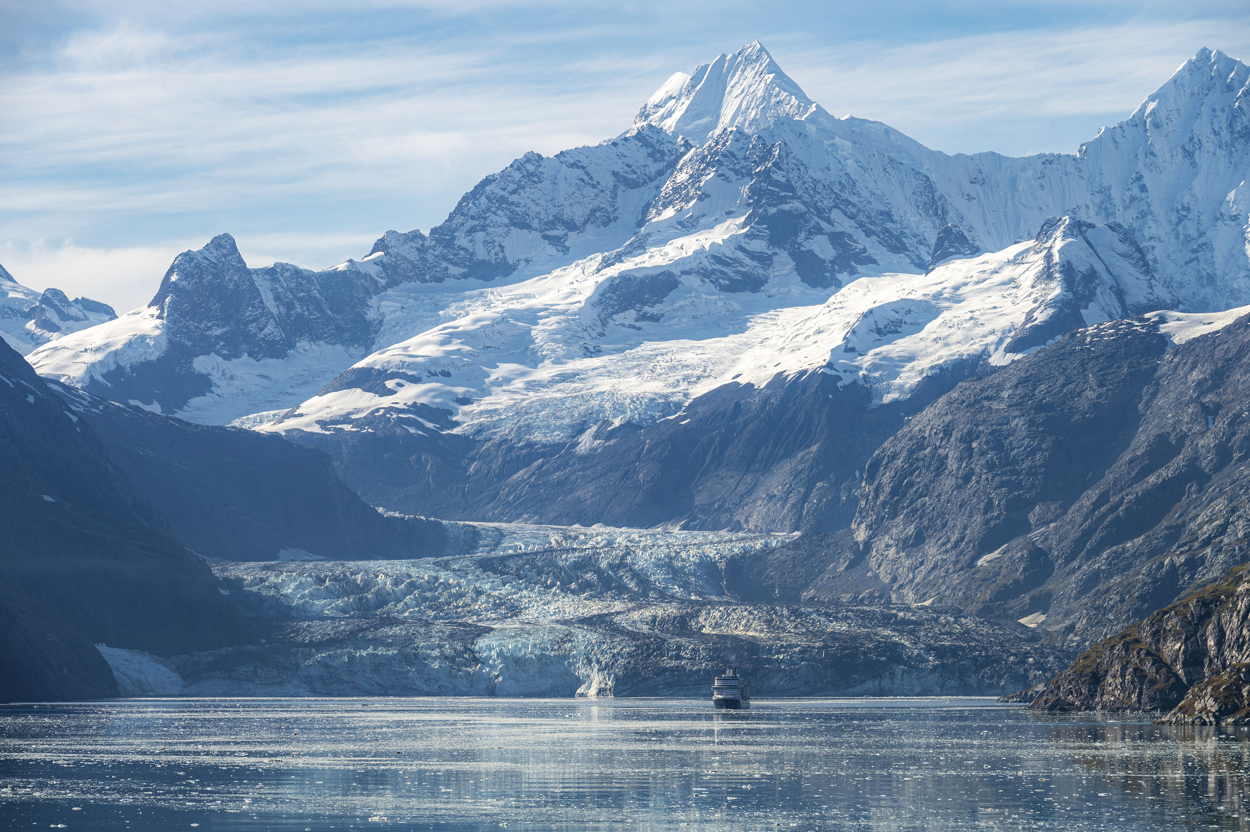 a scenic view of a glacier filling a valley between steep mountains. In the water in front of the glacier, a cruise ship. A towering snowcapped mountain rises to the top of the scene.