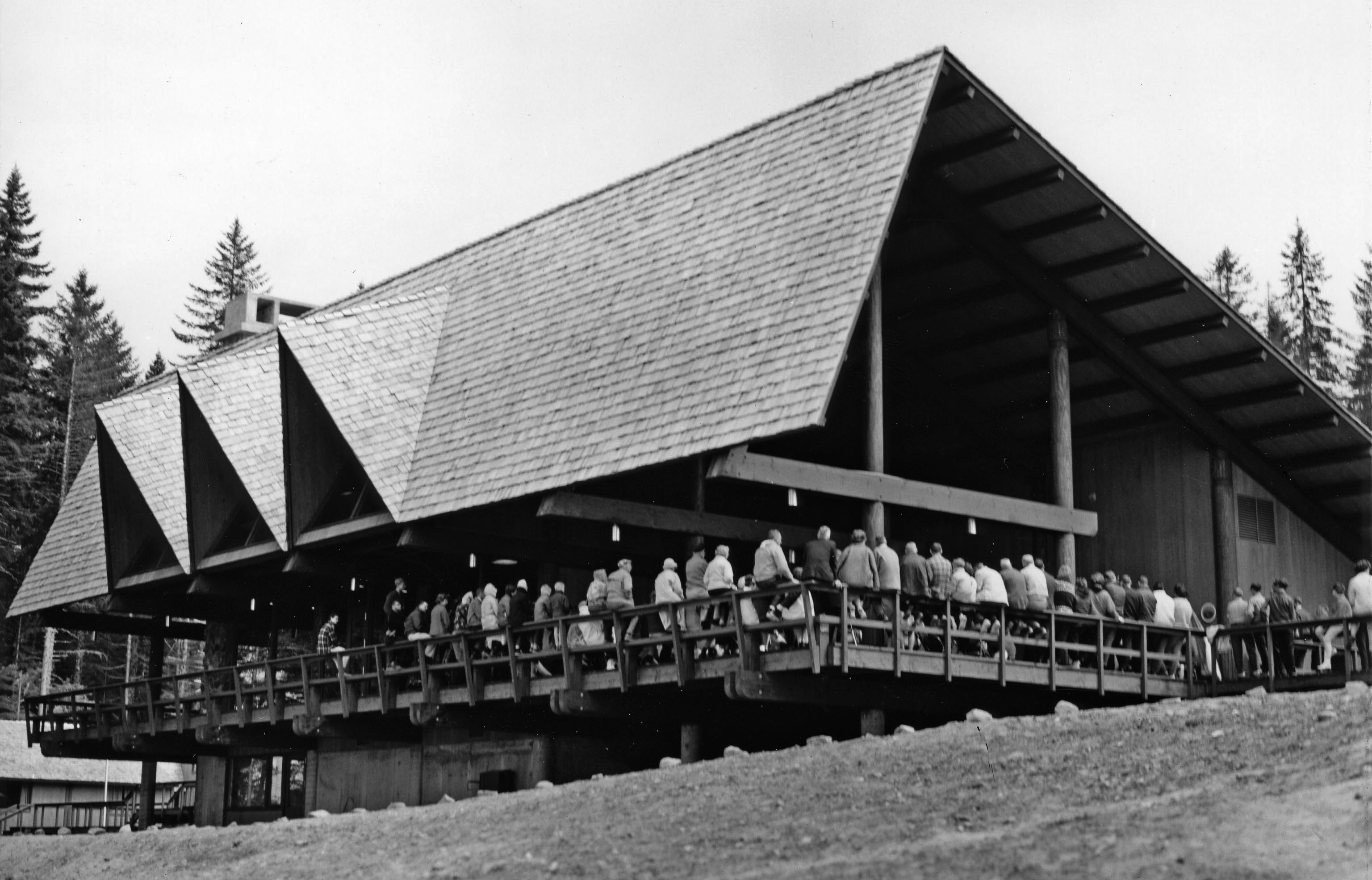 Historic black and white image of the Glacier Bay Lodge from the north side looking south. Many people line the railings at a dedication ceremony