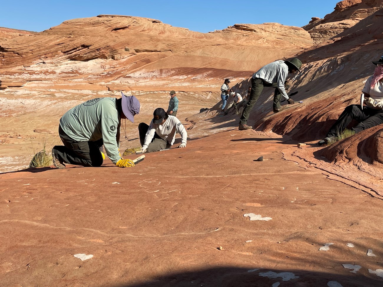 Seven people removing graffiti craved into sandstone with wire brushes, rock hammers and paint scrapers.