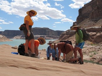 Group of people in desert hiking gear clean sloping slick rock with Lake Powell in background