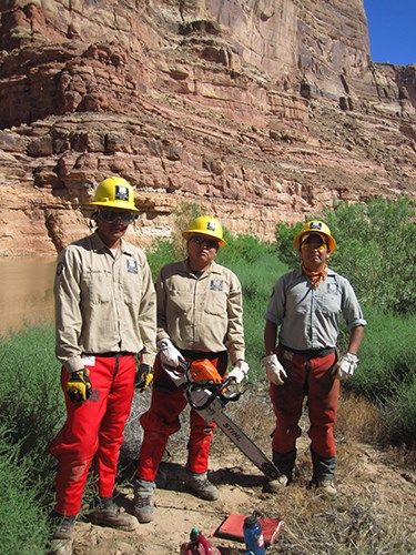 Three SCC participants in hardhats stand on riverbank. One holds a chainsaw