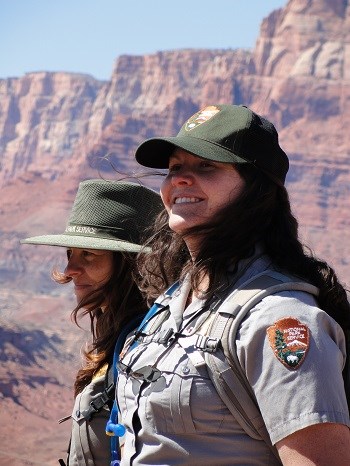 Two uniformed park rangers stand with red cliffs in distance. Their hair blows in the wind.