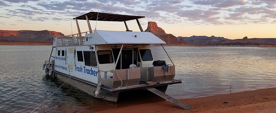 White houseboat labeled Trash Tracker beached during sunset.
