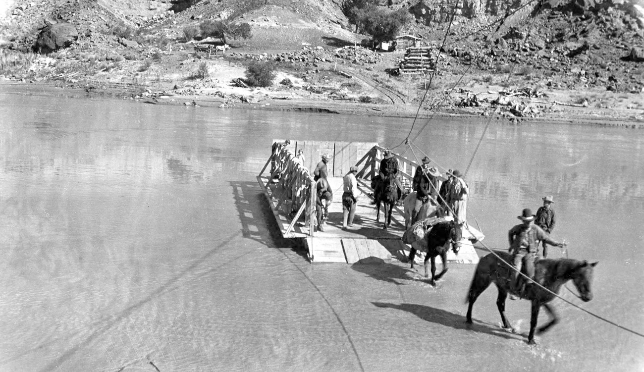 a group of people, some on horseback, exiting a wooden raft at one shore of the river. Cable lines link the shore to the raft and the other side.