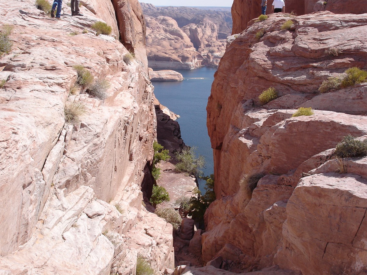People stand on the canyon rim looking down steep rocky incline to Lake Powell below