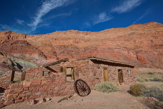 Stacked sandstone bricks of varying sizes make up the walls of a deteriorating building with a log beamed roof. A wagon wheel leans again out front. A tall canyon wall surrounds it in the background..