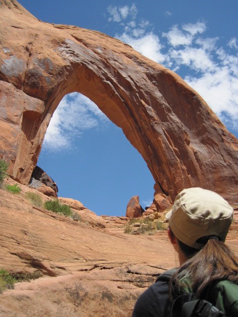 A hiker in a ball cap looks at a tall sandstone arch on a clear day.