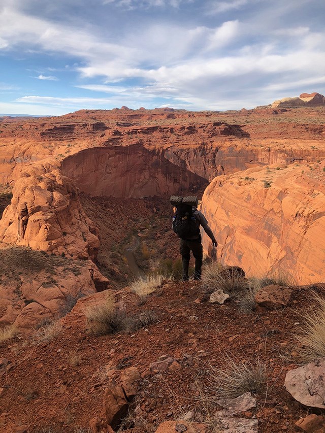 person wearing large backback looks over the rim of a sandstone canyon to a bend in the river at the bottom