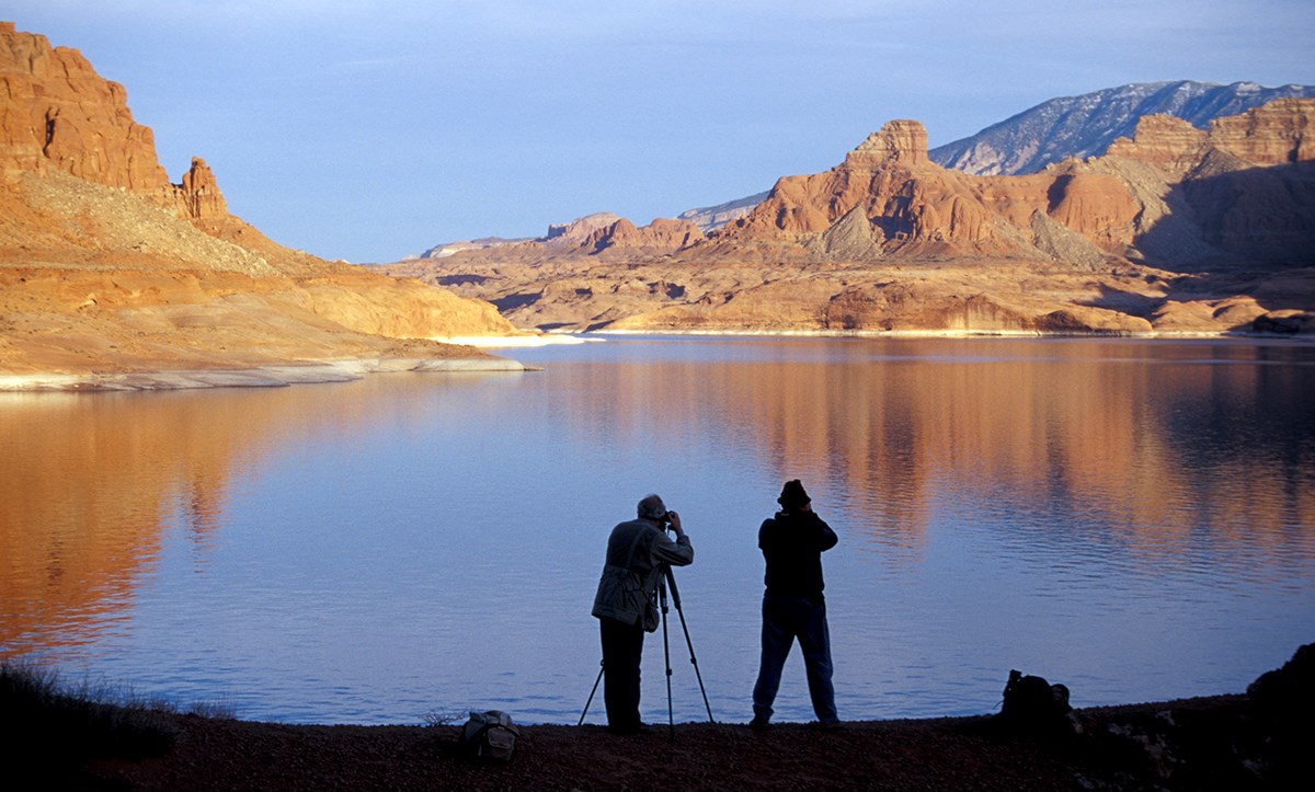 Two photographers in silhouette take photos of sunlit buttes reflected in lake