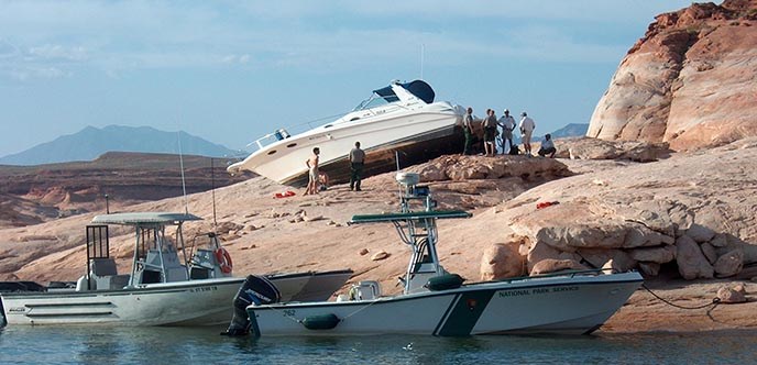 A white boat is stranded on the rocks high above the water. Park Rangers and Sheriffs and their boats surround the area.
