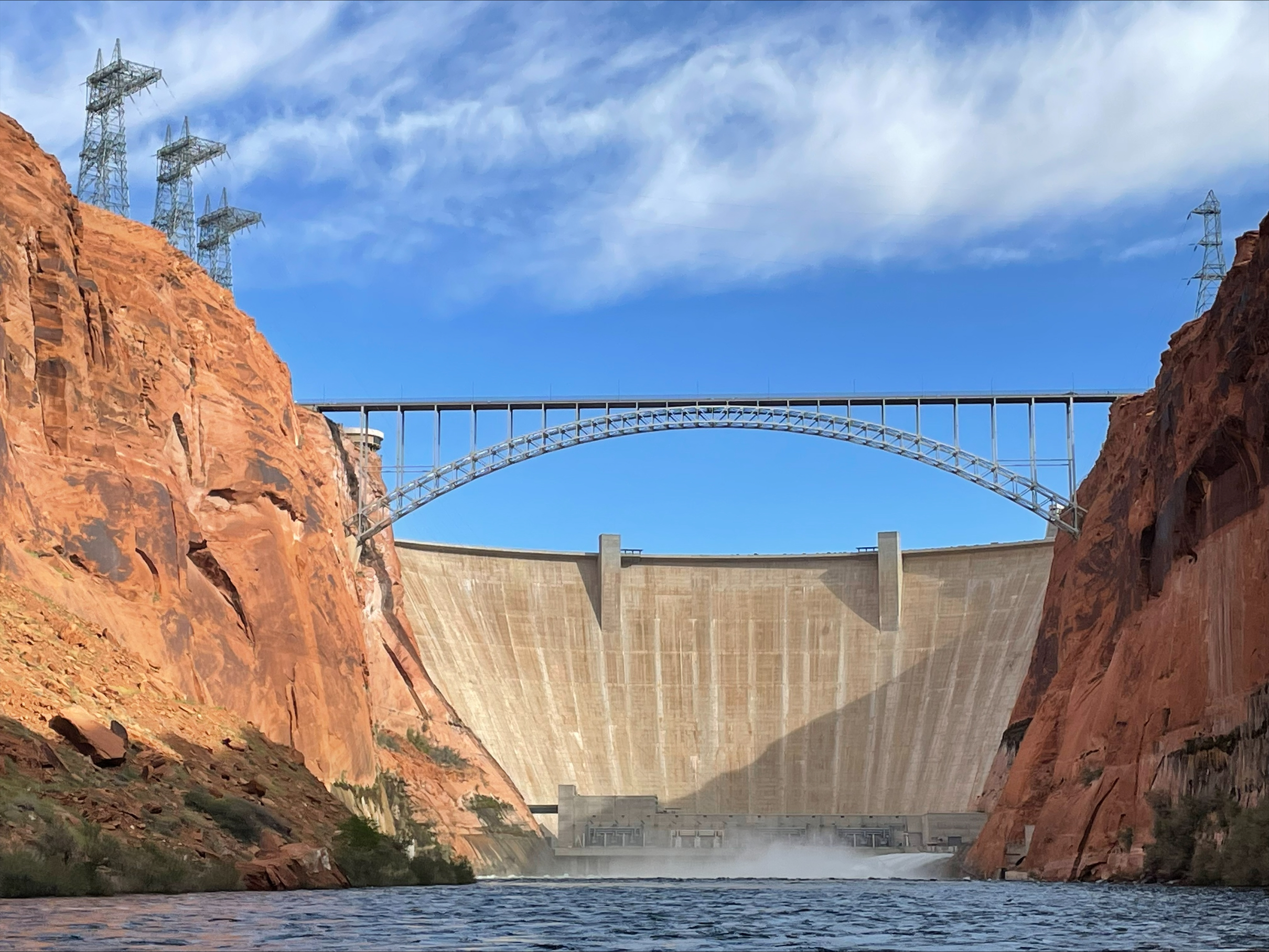 Concrete arch dam in a sandstone canyon. Water rushes out the bottom right side. Steel-arch bridge spans the canyon above the dam.