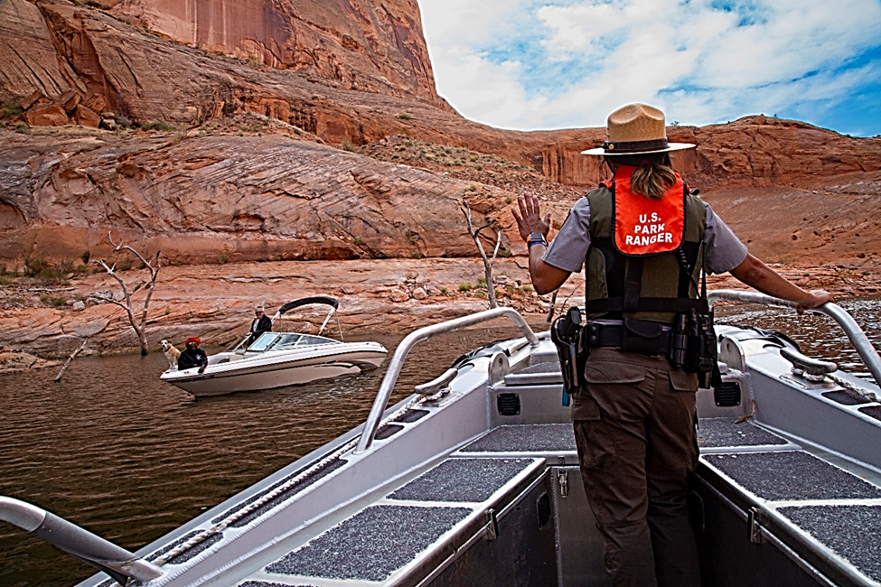 A Park Ranger in a silver boat waves at another boat in the water, surrounded by a redrock canyon.