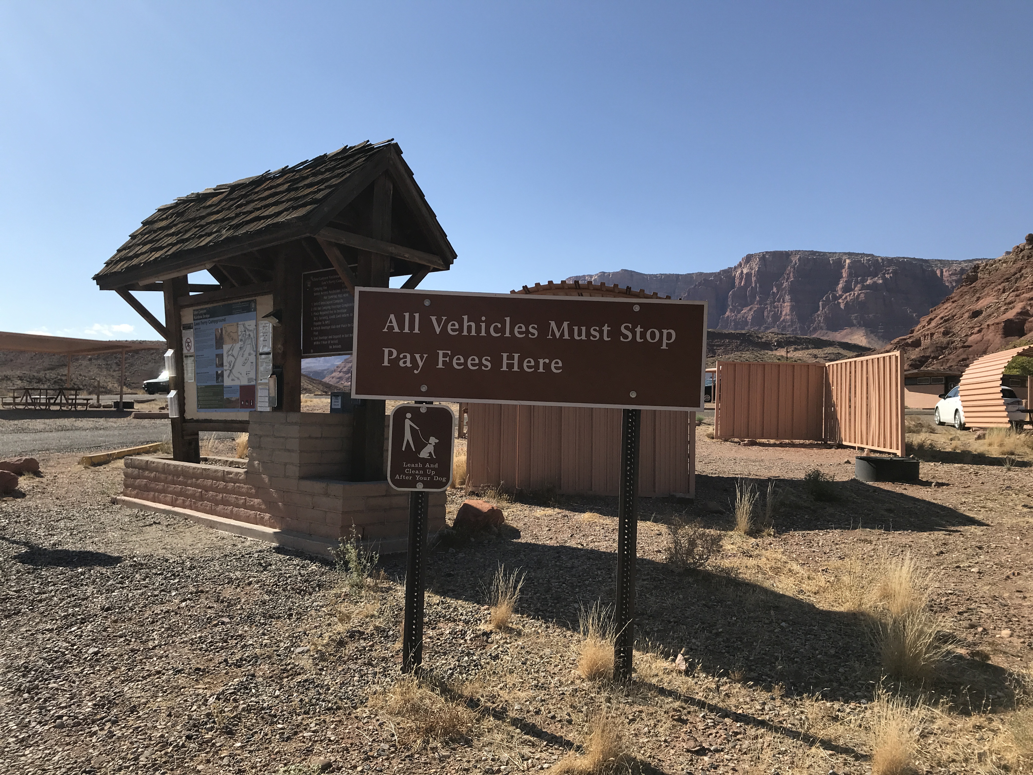A kiosk with a thatched roof has bulletin boards on it. In front of he kiosk is a sign, "All Vehicles Must Stop Pay Fees Here"