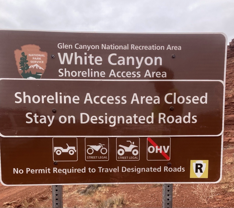 Brown sign with white text and icons of a vehicle, motorcycle, four wheel vehicle, and no off-highway. Words: Glen Canyon National Recreation Area White Canyon Shoreline Access Area Shoreline Access Area Closed Stay on Designated Roads No Permit is requir