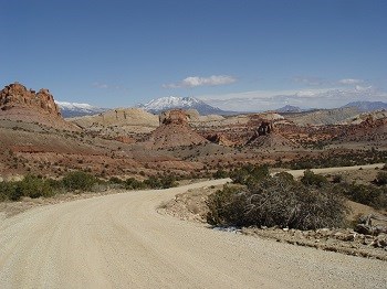Dirt road winds past colorful buttes with snowy mountain in distance