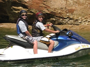 Two boys seated on personal watercraft wearing helmets and life jackets