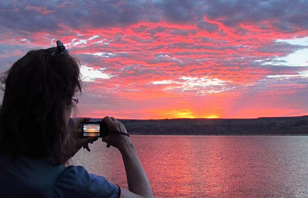 Woman photographs vibrantly colored clouds over lake. The image is repeated in her camera viewscreen.