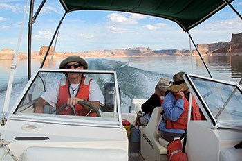 Man wearing life jacket, hat, and sunglasses drives powerboat over Lake Powell with seated passengers also wearing life jackets