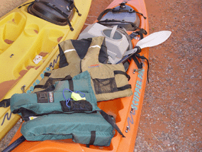 Two kayaks on the ground with the following objects on top of them: two lifejackets, safety whistle, portable toilet, paddle.
