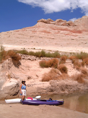 A woman stands on a beach in front of a cliff next to two kayaks.