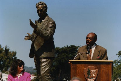 man speaking at podium with statue in background