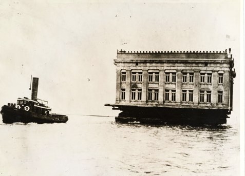 a tug boat pulling a building floating on a barge