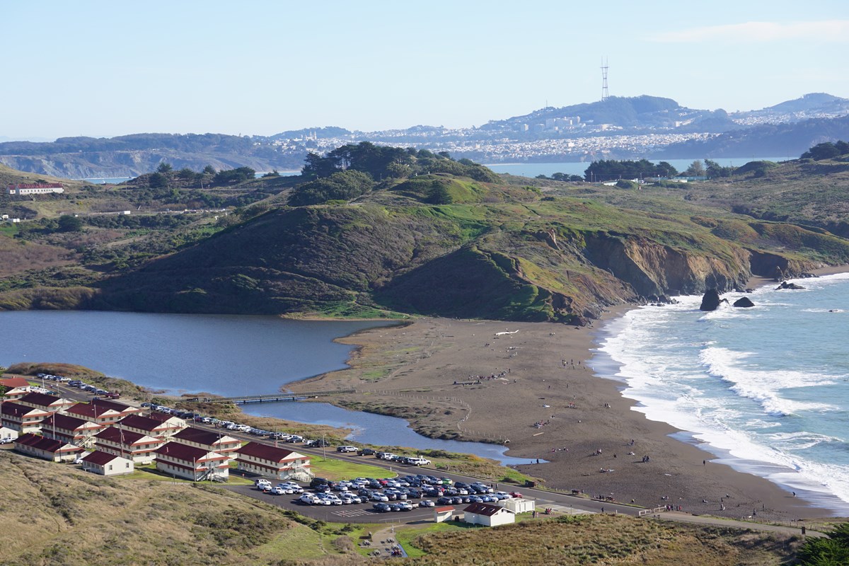 Photo overlooks the beach, with brown sand, bordered on either side by the ocean and the lagoon. A parking lot is near the beach, and the red and white buildings of Fort Cronkhite are across the street from the beach.