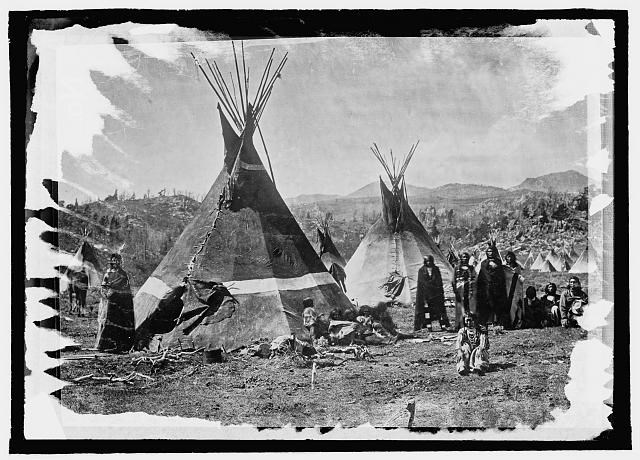 Indigenous Shoshone people stand in front of skin tepees.