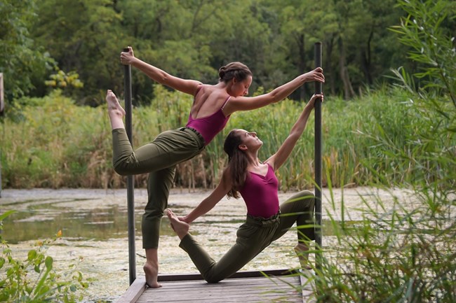 Two dancers hold a pose on a small dock on a river. One is on the ground knees bent and legs going in different directions, while the other hovers above, outstretched and on one foot. They wear matching outfits of pink leotards with dark green pants.