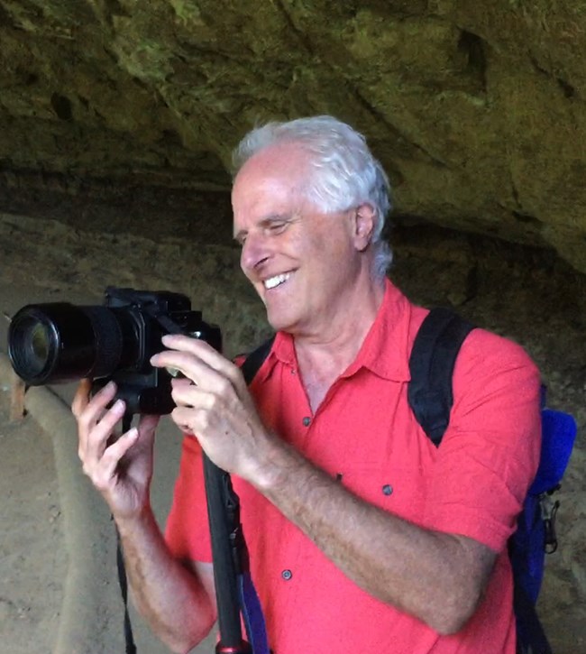 A thinly white haired man in a bright, salmon colored shirt holds a camera with a big smile on his face. He stands in front of a rock wall, looking like a cave or cliff dwelling.