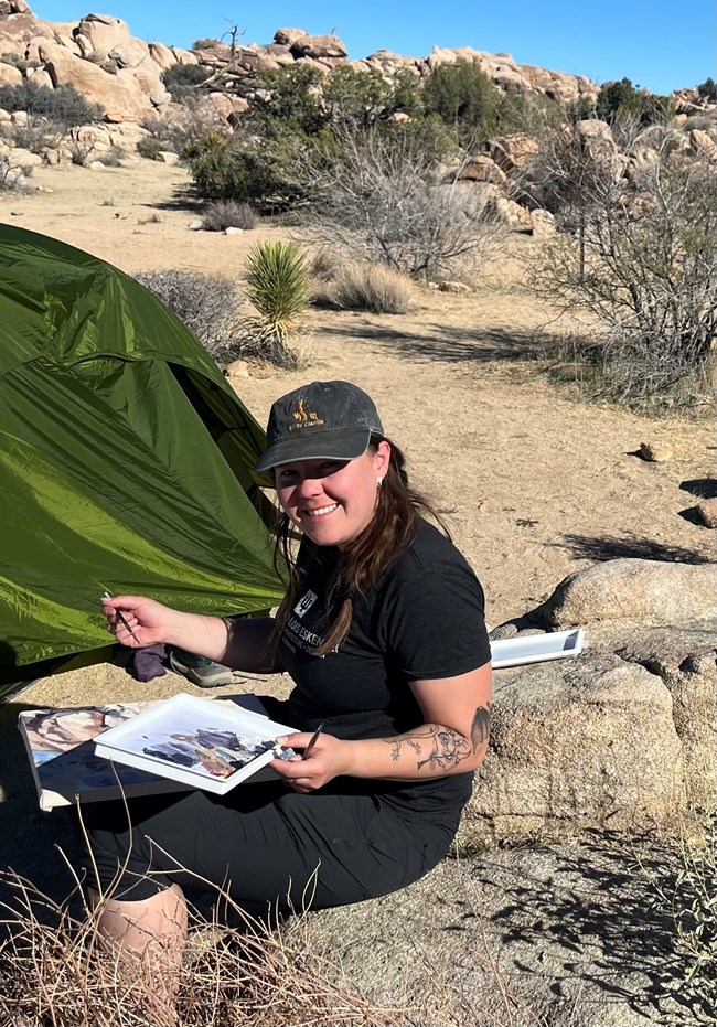 An artist wearing all black with tattoos on her left arm smiles with her palette and paintbrush beside a tent amid a wild landscape of granite stones and desert plants.